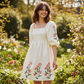 Beige floral print puff sleeve babydoll dress with pink flower embroidery, worn by a smiling woman in a garden.
